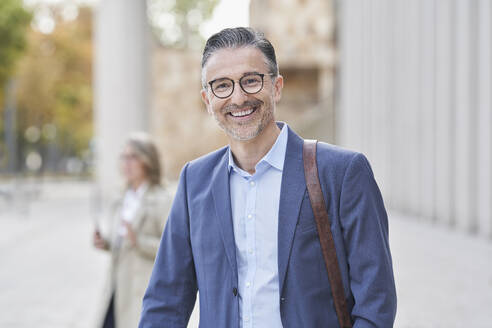 Happy mature businessman in blazer standing on footpath - RORF03286