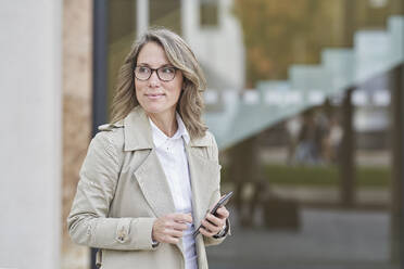 Nachdenkliche reife Geschäftsfrau steht mit Smartphone vor einem Gebäude - RORF03293