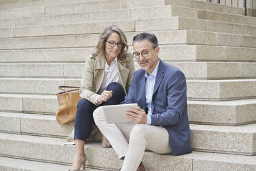 Mature businessman using tablet PC sitting by colleague on steps - RORF03309