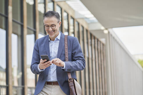 Happy mature businessman using mobile phone outside building - RORF03315