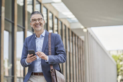 Smiling mature businessman holding mobile phone outside building - RORF03316