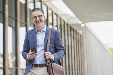 Happy mature businessman standing with smart phone outside building - RORF03317