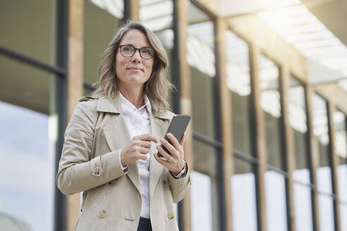 Contemplative mature businesswoman standing with mobile phone outside building - RORF03322