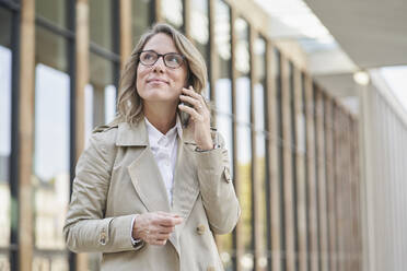 Nachdenkliche reife Geschäftsfrau, die vor einem Gebäude über ihr Smartphone spricht - RORF03326