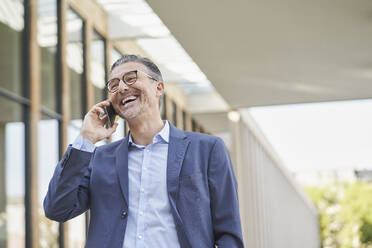 Glücklicher Geschäftsmann, der vor einem Gebäude mit seinem Smartphone telefoniert - RORF03329