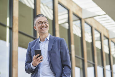 Cheerful mature businessman with mobile phone standing outside building - RORF03331