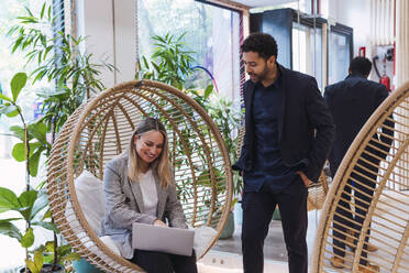 Businesswoman sitting with laptop in hanging chair talking to businessman - PNAF04670