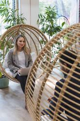 Businesswoman sitting with laptop in hanging chair talking to businessman - PNAF04673