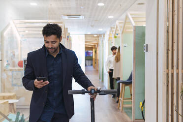 Geschäftsmann mit Elektroroller und Mobiltelefon im Büro - PNAF04676