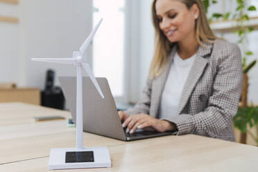 Young businesswoman using laptop and digital tablet at desk with wind turbine model in foreground - PNAF04762