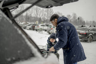 Vater mit Sohn sitzt im Einkaufswagen auf verschneitem Parkplatz - ANAF00778