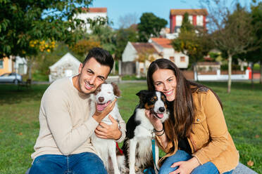 Husband and wife embracing purebred dogs while sitting on bench near grassy lawn against residential buildings in daylight and looking at camera - ADSF42169