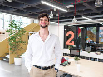 Smiling young bearded man in smart casual outfit standing near table with laptop in modern spacious workspace and looking at camera - ADSF42179