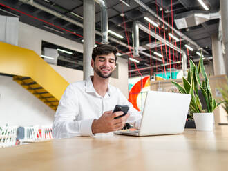 Glücklicher junger bärtiger Mann in weißem Hemd sitzt am Tisch und surft mit seinem Smartphone im Internet, während er an seinem Laptop in einer geräumigen Büro-Cafeteria arbeitet - ADSF42181