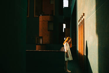 Side view of redhead lady in white dress standing in dark hallway of contemporary building in Barcelona while sun shining through window looking down - ADSF42196
