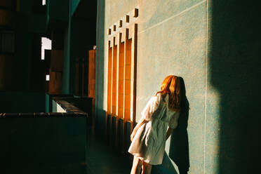 Back view of unrecognizable redhead lady in white dress standing in dark hallway of contemporary building in Barcelona while sun shining through window - ADSF42197