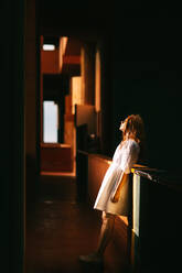 Side view full body of young female in white dress standing in dark hallway against window with sunlight in Walden 7 building - ADSF42202