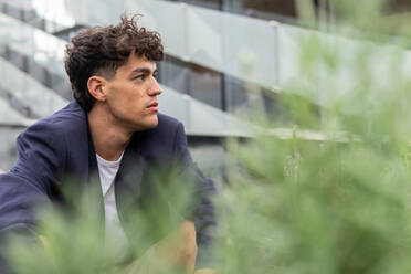 Young male in smart casual clothes sitting on haunches behind green shrub and looking away while spending time on terrace of high rise building - ADSF42236