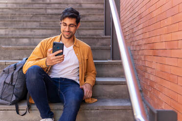 Fokussierter junger bärtiger Hispanoamerikaner in Jeans und mit Brille, der auf einer Steintreppe mit Rucksack sitzt und tagsüber mit dem Handy im Internet surft - ADSF42240