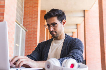 Konzentrierter bärtiger männlicher Student mit Brille und Freizeitkleidung, der an einem Holztisch in der Nähe eines Headsets sitzt und Informationen auf einem Laptop in einem modernen Arbeitsraum mit Backsteinsäulen eintippt - ADSF42242