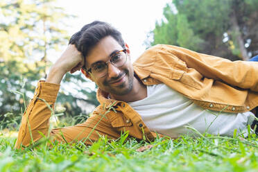 Smiling bearded ethnic man in eyeglasses lying on green grass with yellow leaves and looking at camera - ADSF42245