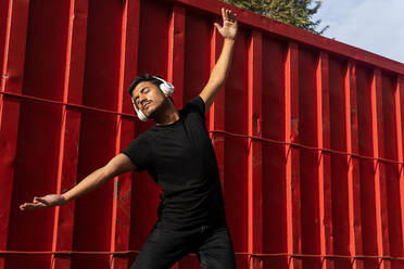 Energetic man in black outfit dancing while listening to favorite song on headphones against red metal fence - ADSF42246
