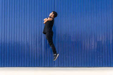 Side view full body of active male dancer jumping on street while embracing shoulders against blue striped wall - ADSF42249