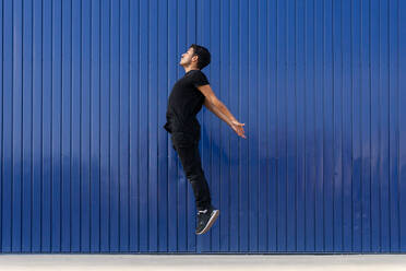 Side view full body of active male dancer jumping on street against blue striped wall - ADSF42250