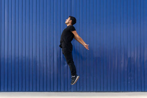Seitenansicht Ganzkörperansicht eines aktiven männlichen Tänzers, der auf der Straße gegen eine blau gestreifte Wand springt - ADSF42250