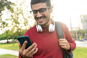 Smiling young man in red sweatshirt and headphones on neck with rucksack standing on city street browsing on mobile phone - ADSF42253