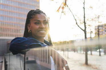 Seitenansicht einer positiven afroamerikanischen Frau mit Zöpfen, die auf der Straße vor einem modernen Gebäude sitzt, lächelt und wegschaut - ADSF42254