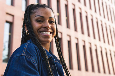 Low angle of positive African American female with braids standing on street against contemporary building smiling and looking at camera - ADSF42255