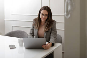 Adult female freelancer with long brown hair in casual wear working remotely on laptop sitting at table at home - ADSF42260
