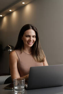 Selbstbewusste Freiberuflerin mittleren Alters mit langen braunen Haaren in Freizeitkleidung lächelnd beim Tippen auf dem Laptop während der Online-Arbeit zu Hause - ADSF42261
