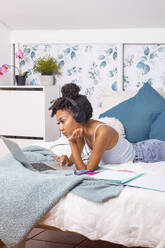 Spanische Teenagerin in Freizeitkleidung und mit Kopfhörern, die zu Hause auf dem Bett liegend mit einem Laptop lernt - ADSF42275