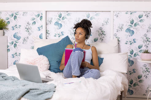 Spanische Teenagerin in Freizeitkleidung und mit Kopfhörern, die in ein Notizbuch schreibt, während sie zu Hause auf dem Bett neben einem Laptop sitzt - ADSF42276