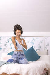 Fröhliche hispanische Teenagerin in Freizeitkleidung und Kopfhörern, die Musik hört, während sie auf dem Bett zu Hause tanzt und in die Kamera schaut - ADSF42277