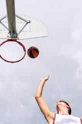 From below of anonymous sportive male throwing ball into basketball hoop while playing game against blue sky on sunny summer day - ADSF42278