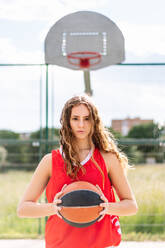 Frau in Sportkleidung mit Ball steht auf einem Sportplatz im Freien und blickt in die Kamera gegen ein Brett mit einem Basketballkorb - ADSF42279