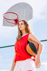 From below empowered young woman in activewear with ball standing on outdoor sports ground and looking away against backboard with hoop - ADSF42280