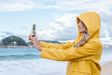 Seitenansicht einer Frau mit langen blonden Haaren in einem gelben Regenmantel, die ein Selfie mit ihrem Smartphone auf einem unscharfen Hintergrund einer grünen Insel in Spanien macht - ADSF42285