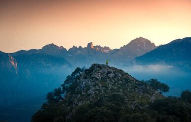 Picturesque scenery of mountainous valley covered with fog and anonymous tourist standing on edge under bright sunset sky in Asturias - ADSF42307