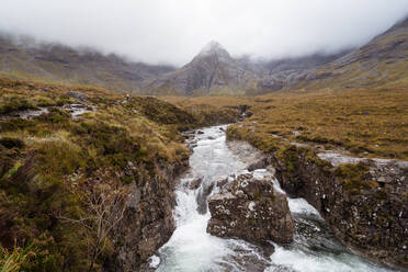 Malerischer Blick auf eine mit Gras bewachsene Bergkette und einen fließenden Bach unter nebligem Himmel in Schottland - ADSF42317