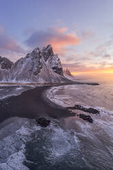 Calm frozen endless sea surrounding with black frozen sand and high rocky snowy Vestrahorn mountain during picturesque sunset in Stockness beach, Iceland - ADSF42332