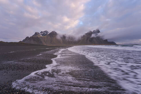 Ruhiges, gefrorenes, endloses Meer, umgeben von schwarzem, gefrorenem Sand und dem hohen, felsigen, schneebedeckten Berg Vestrahorn in Stockness Beach, Island - ADSF42333