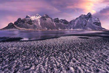 Ruhiges, gefrorenes, endloses Meer, umgeben von schwarzem, gefrorenem Sand und dem hohen, felsigen, schneebedeckten Berg Vestrahorn bei malerischem Sonnenuntergang am Strand von Stockness, Island - ADSF42335