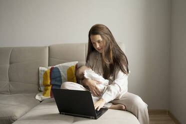 Young mom embracing infant and browsing data on laptop while sitting on couch and working on remote project at home - ADSF42337