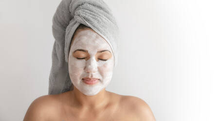 Fröhliche junge Frau mit Handtuch auf dem Kopf, die sich einer Hautpflegebehandlung unterzieht, während sie vor einem weißen Hintergrund steht - ADSF42342