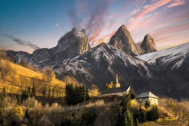 Picturesque view of small church building located on hill near mountain range covered with snow under colorful sky in evening - ADSF42345