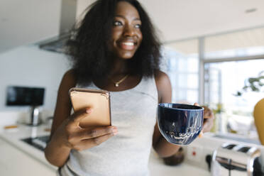 Smiling woman holding smart phone and tea cup at home - TYF00555